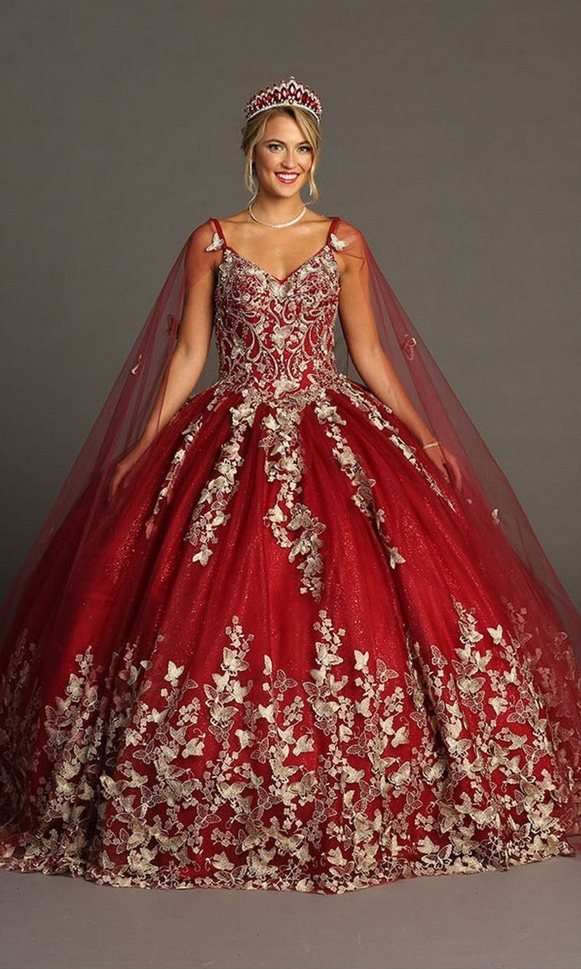 A woman stands against a gray background wearing the Quinceanera Dress CHF3077 By Chicas, a striking red gown adorned with gold floral embroidery and delicate butterfly appliques. She completes the elegant look with a matching cape and jeweled crown.